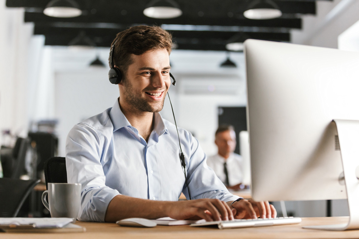 Photo of businesslike man 20s wearing office clothes and headset working on computer in call center