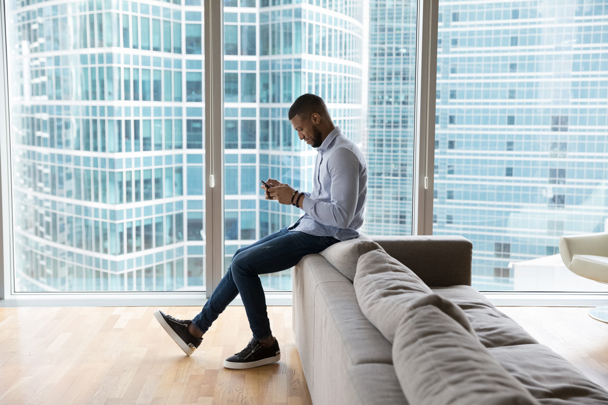Positive successful young African business man typing on mobile phone in office lobby, hotel room, in urban home apartment, chatting, using online app, sitting against city view from big window