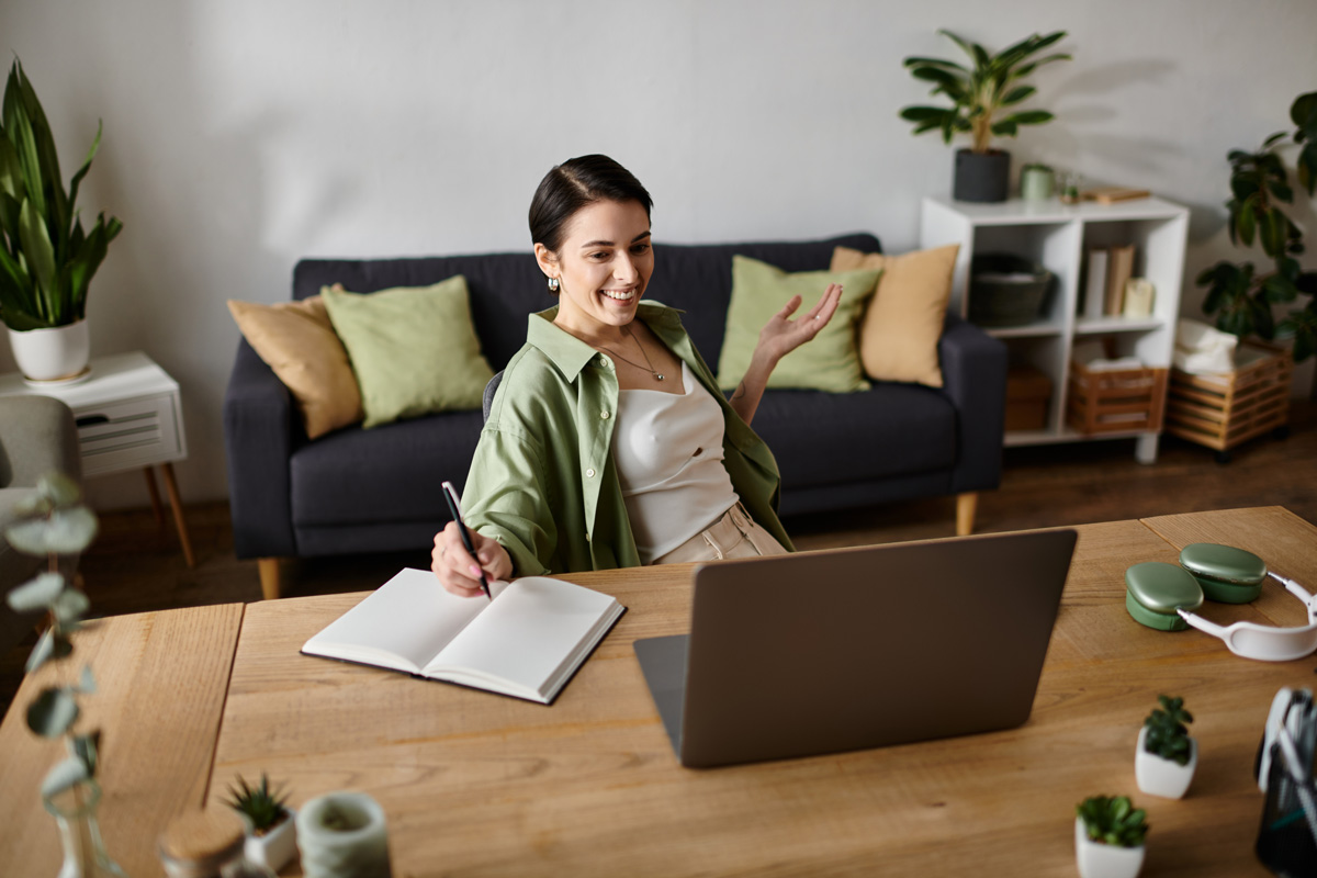 A woman smiles while working from home, sitting at a desk with a laptop and a notebook for her home-based-business.